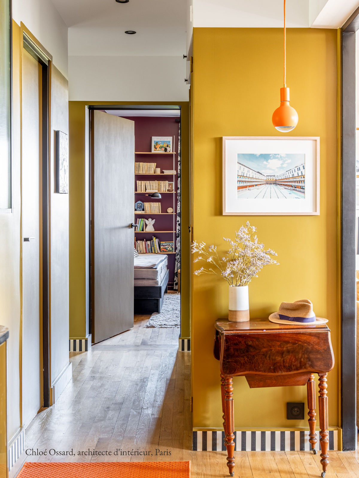 Couloir aux murs jaunes avec la frise Tempo Noir & Écru posée en plinthe au ras du parquet, petite table en bois et suspension orange, par Chloé Ossard